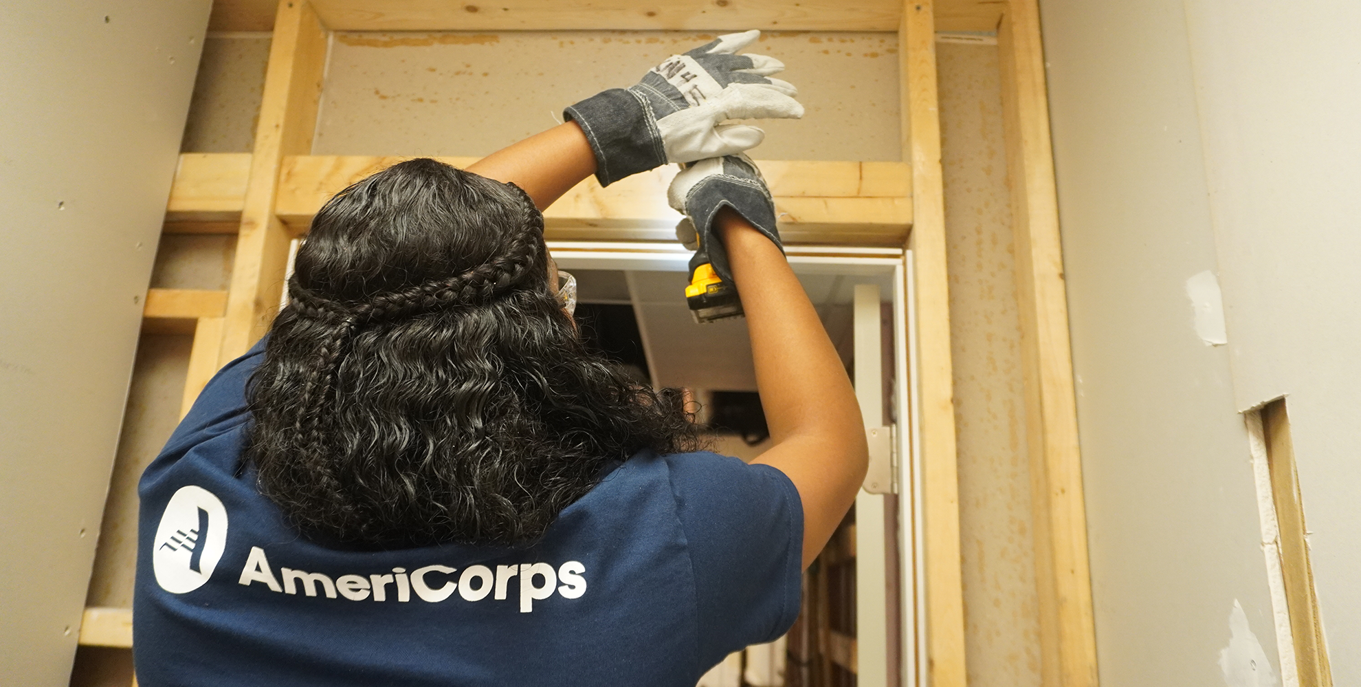 Woman wearing an AmeriCorps t-shirt drills into a wall.