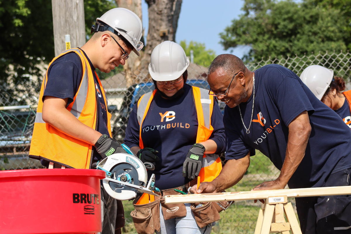 Three people working together outside, standing over a piece of wood with a rotary saw