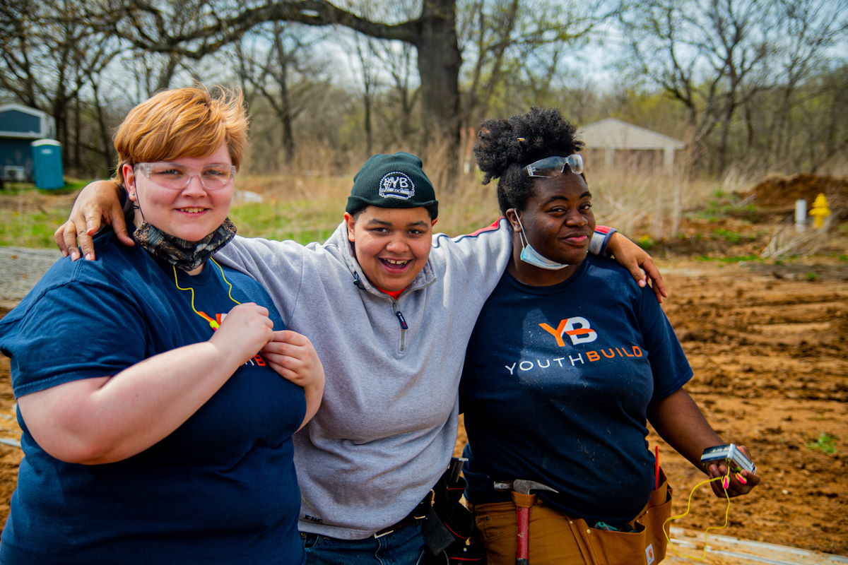 Three people standing together outside, arms around each other's shoulders and smiling